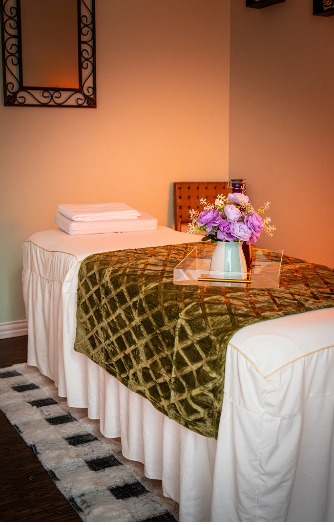 Spa treatment room with two massage tables draped in white linens and gold patterned covering, decorative purple flowers in a vase, and warm peach-colored walls