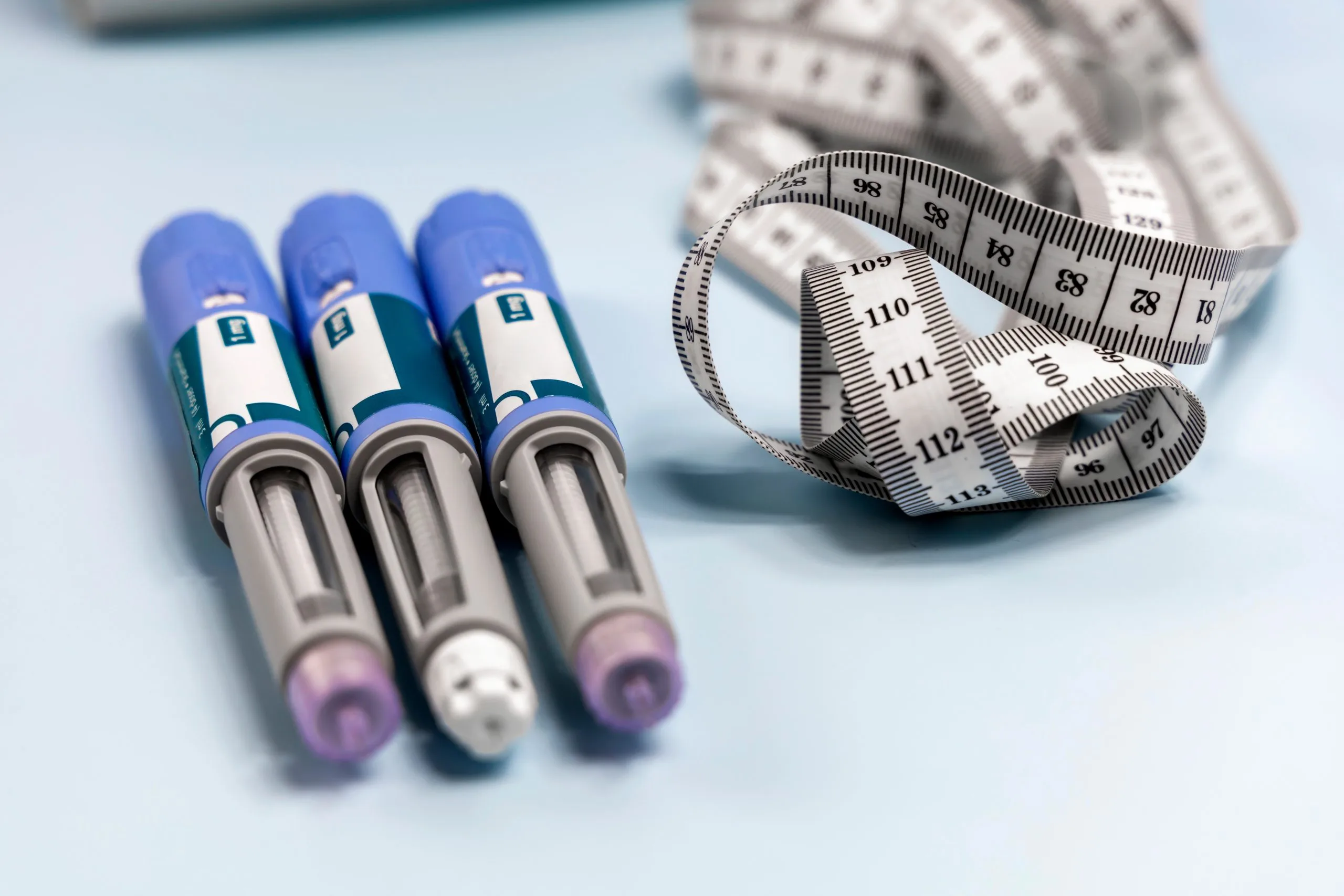 Three pre-filled semaglutide injection pens next to a measuring tape on a light blue surface